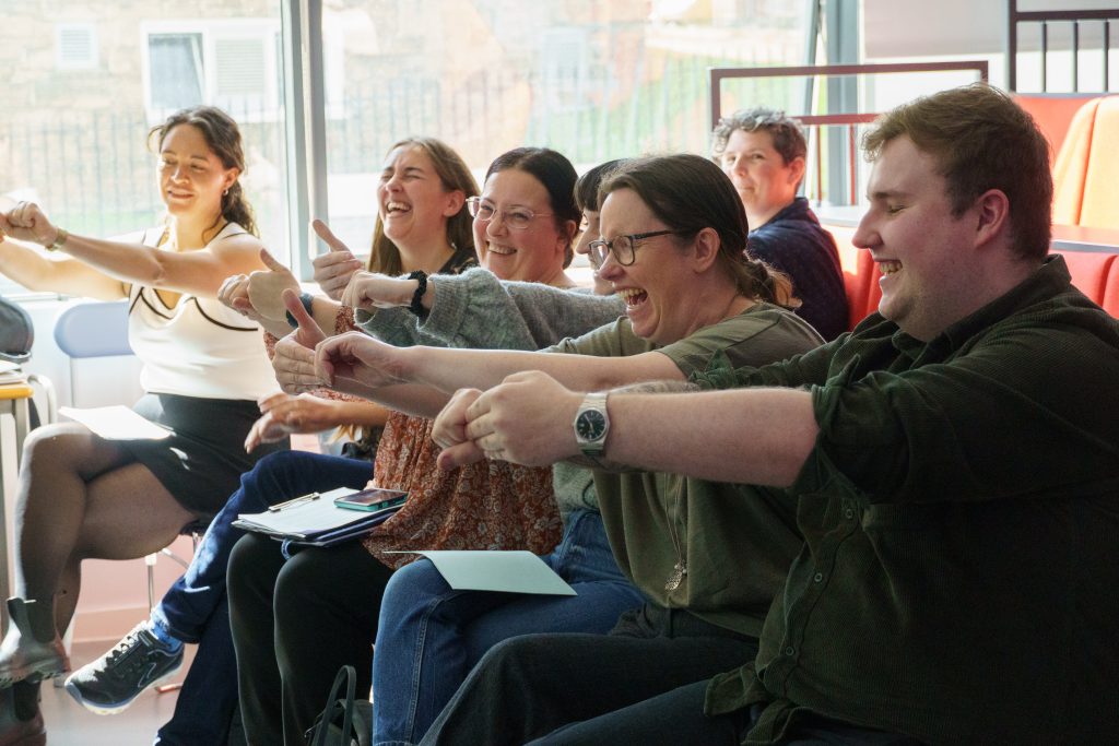A group of adults sitting indoors, laughing and giving thumbs-up gestures together during a lively group activity.